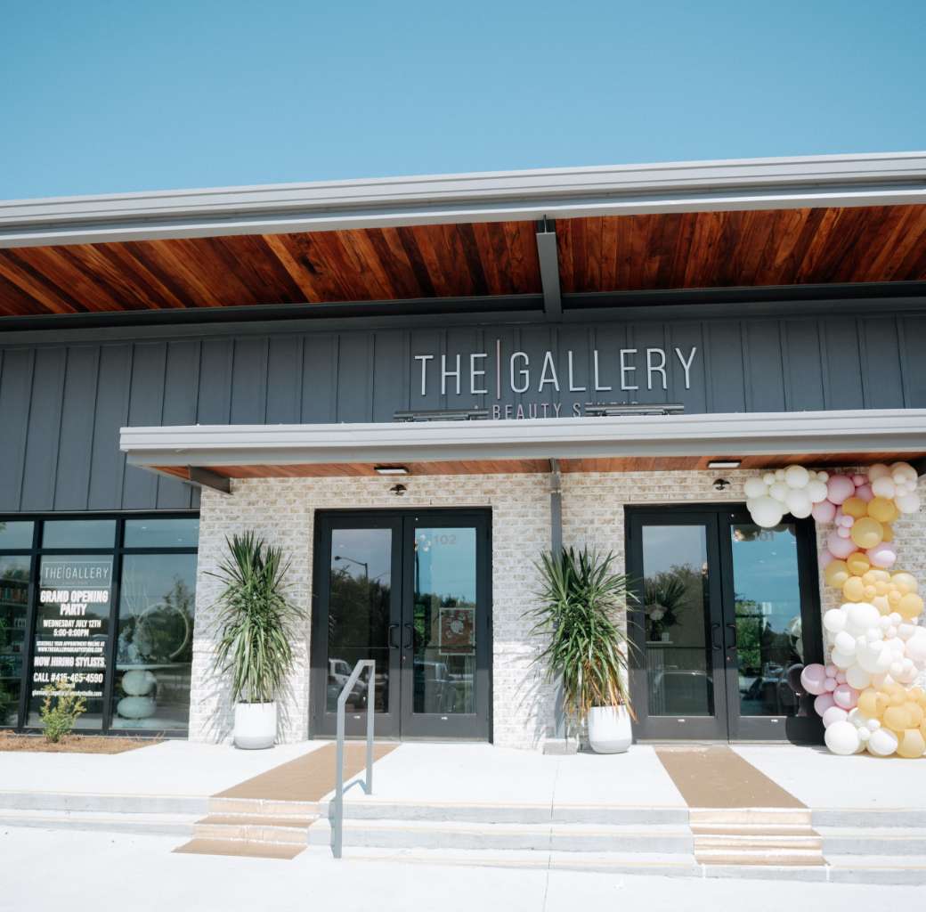 The Gallery A Beauty Studio - The exterior of The Gallery Beauty Salon, a full service hair salon in Charleston, features large glass doors, potted plants, cream brick walls, and pastel balloons decorating the entrance under a clear blue sky.