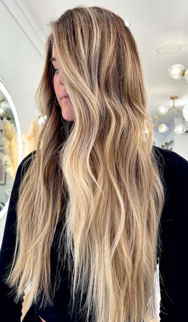 The Gallery A Beauty Studio - A woman with long, wavy, blonde hair featuring darker roots and lighter highlights stands indoors at a Charleston Hair Salon, wearing a black top. The softly lit background showcases modern light fixtures.