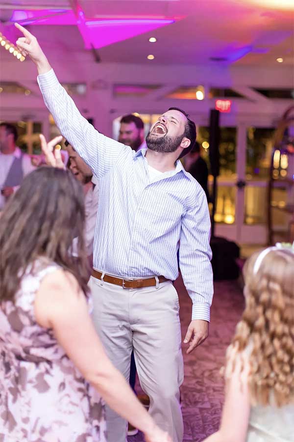 Bunn DJ Company - A man in a striped shirt dances enthusiastically at an indoor event, raising one arm and singing, while others dance around him.