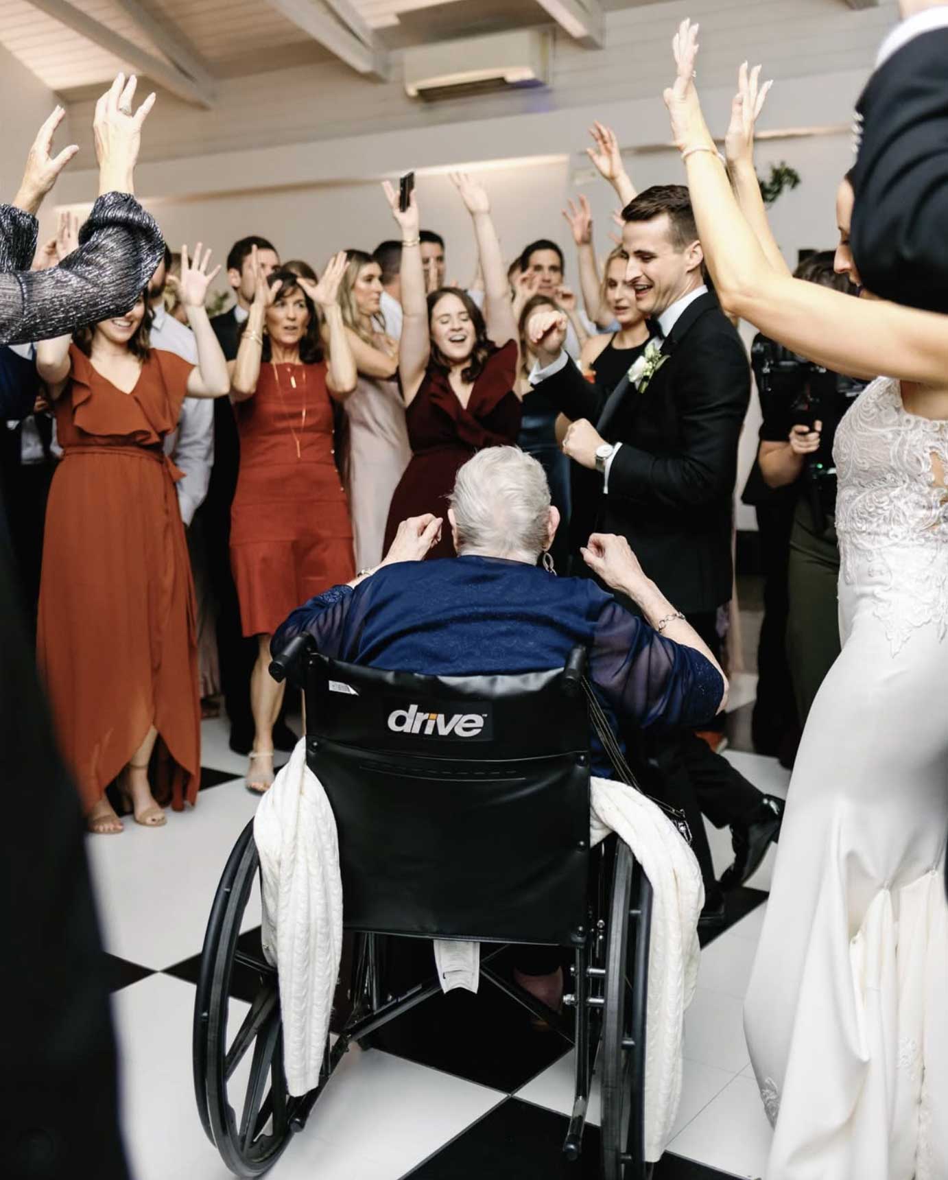 Bunn DJ Company - An older woman in a wheelchair faces a dancing group at an indoor event, with people around her raising their hands and celebrating on a checkered floor.